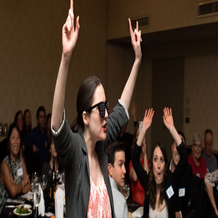 Woman with sunglasses and blazer raises her arms at a crowded banquet, others cheering around a long table.